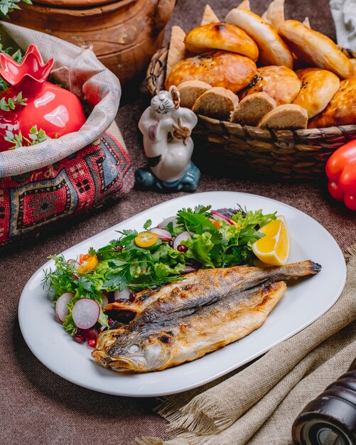 Vista lateral de pescado a la parrilla con una ensalada de verduras y hierbas con una rodaja de limón