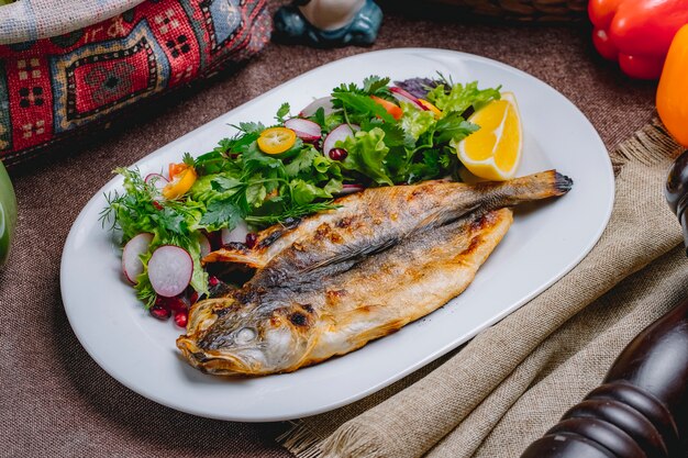 Vista lateral de pescado a la parrilla con una ensalada de verduras y hierbas con una rodaja de limón