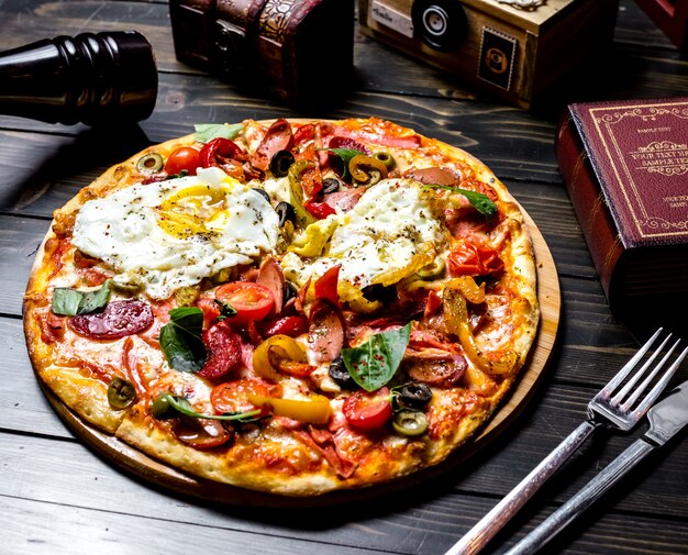 Vista lateral pizza mixta con tomates aceitunas pimiento huevos salchichas en el tablero un libro un cuchillo y un tenedor sobre la mesa