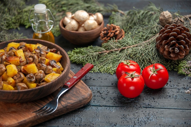 Vista lateral del plato de comida tazón de madera de papas con champiñones en la tabla de cortar junto al tenedor y tomates debajo del tazón de aceite de setas blancas y ramas de abeto