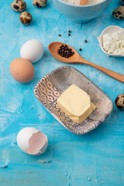 Vista lateral del plato de mantequilla y cáscara de huevo con huevos sobre fondo azul.