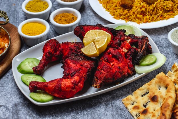 Vista lateral de pollo al horno con pepino, condimento de limón y pan sobre la mesa