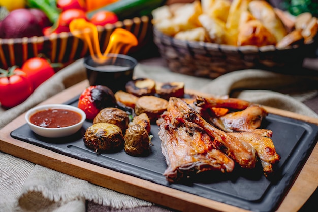 Vista lateral de pollo asado con papas al horno verduras a la parrilla y salsa en un tablero negro