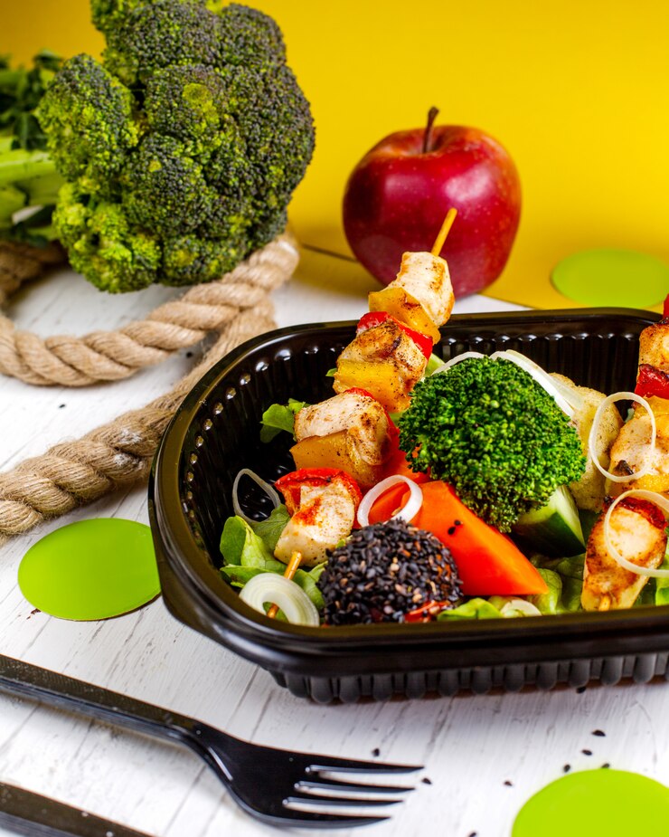 Foto gratuito vista lateral del pollo frito en brochetas con verduras frescas, brócoli y cebolla en una caja de entrega