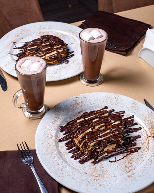 Vista lateral de postre con plátanos cubiertos de chocolate y servido con cacao con malvavisco en vidrio sobre la mesa