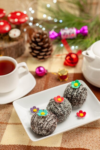 Vista lateral de primer plano galletas galletas y una taza de té en un platillo junto a la tetera y las ramas de los árboles con juguetes de árbol de Navidad en el mantel a cuadros