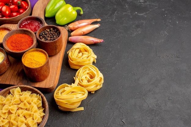 Vista lateral de primer plano pasta y especias tazón de cebolla de tomates pimiento junto a las salsas y especias en la tabla de madera y pasta en la mesa