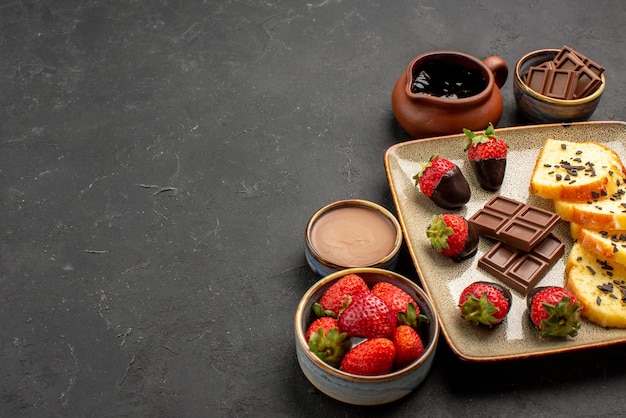 Vista lateral de primer plano pastel de postre con fresas cubiertas de chocolate y chocolate con tazones de crema de chocolate y bayas en el lado derecho de la mesa oscura
