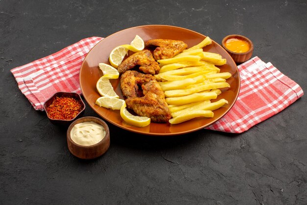 Vista lateral de primer plano plato de comida rápida apetitosas alitas de pollo papas fritas con limón y tazones de salsas y especias sobre un mantel a cuadros rosa-blanco sobre la mesa negra