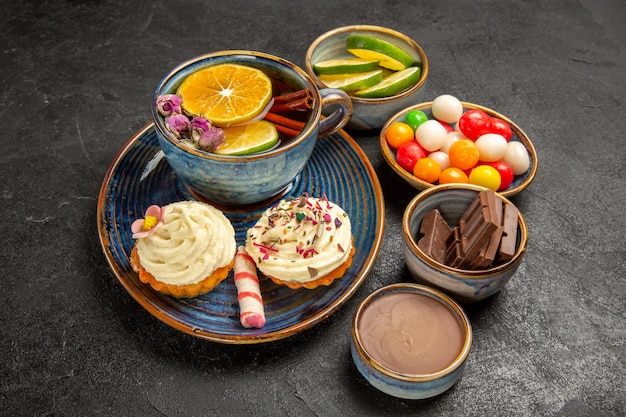 Vista lateral de primer plano té de hierbas una taza de té con canela y limón y los apetitosos cupcakes junto a los cuencos de rebanadas de chocolate de crema de chocolate de lima y dulces coloridos sobre la mesa