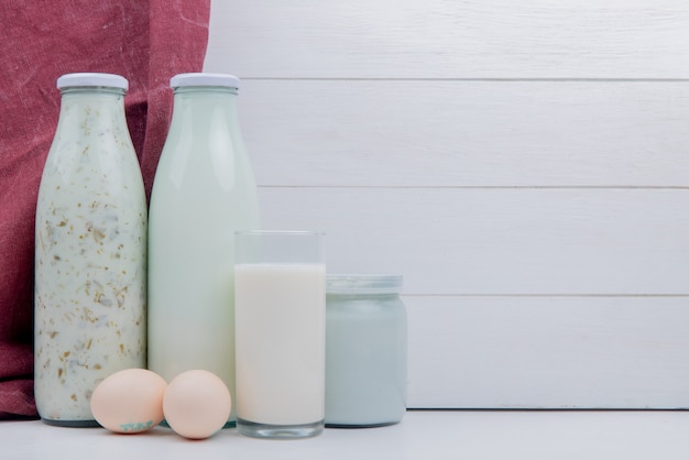 Vista lateral de productos lácteos como leche de yogurt azerbaiyano y leche agria coagulada con huevos en la superficie blanca y mesa de madera con espacio de copia