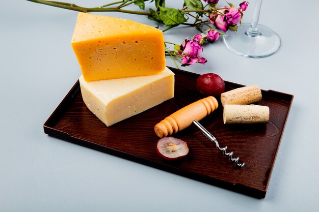 Vista lateral de quesos como queso cheddar y parmesano con corchos de uva y sacacorchos en tabla de cortar y flores en blanco