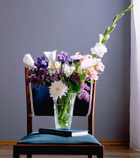 Vista lateral de un ramo de alcatraces de color blanco con iris morado oscuro lila y flores de gladiolo blanco en un florero de vidrio de pie sobre un libro en una silla en el fondo de la pared gris