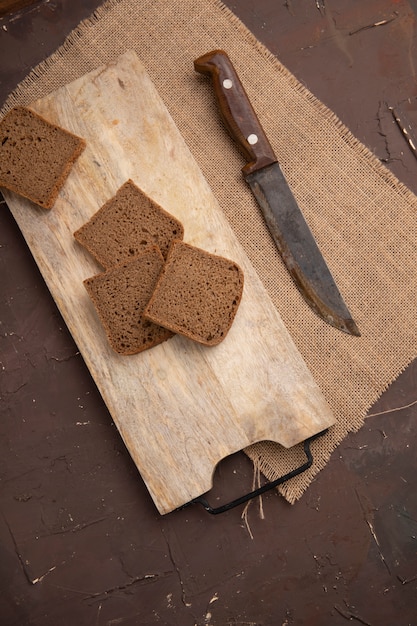 Vista lateral de rebanadas de pan de centeno en la tabla de cortar y un cuchillo en tela de saco sobre fondo marrón con espacio de copia