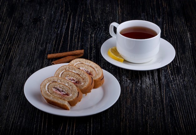 Vista lateral de rebanadas de rollo suizo con crema batida y mermelada de frambuesa en un plato servido con una taza de té en rústica