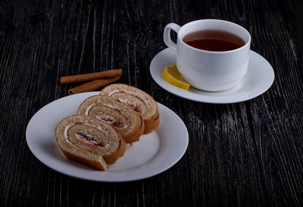 Vista lateral de rebanadas de rollo suizo con crema batida y mermelada de frambuesa en un plato servido con una taza de té en rústica