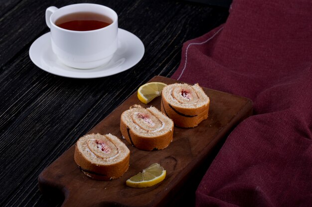 Vista lateral de rebanadas de rollo suizo con mermelada de frambuesa sobre una tabla de madera servida con una taza de té