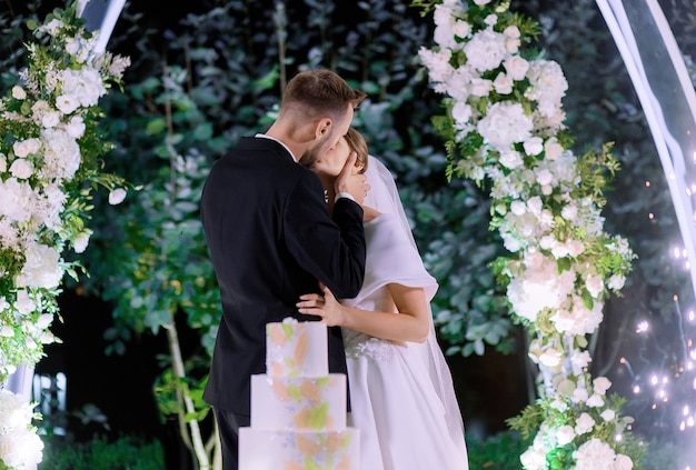 Vista lateral de los recién casados besándose durante la celebración de la boda sobre un fondo de decoración con flores