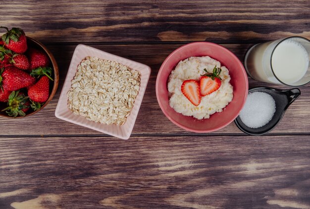 Vista lateral de requesón con fresas frescas en un tazón copos de avena azúcar y vaso de leche en madera rústica