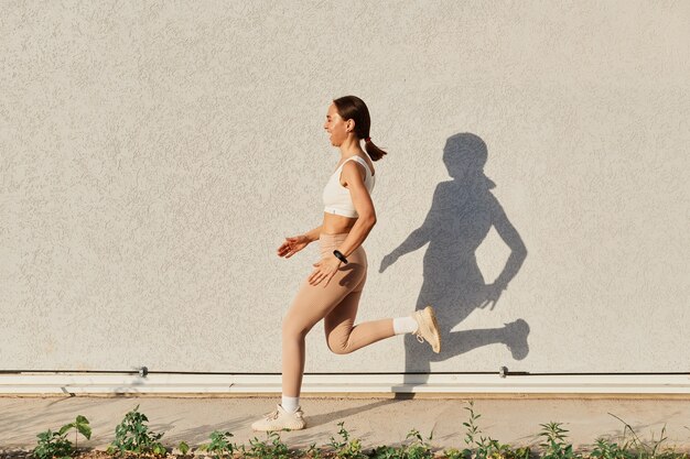 Vista lateral retrato de cuerpo entero de mujeres riendo vistiendo top blanco y leggins beige corriendo afuera