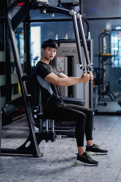 Vista lateral, retrato joven guapo en ropa deportiva sentado para hacer ejercicio de prensa de pecho de máquina en el gimnasio moderno,