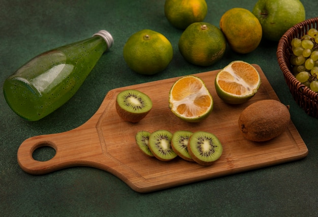 Foto gratuita vista lateral de rodajas de kiwi sobre una tabla de cortar con mandarinas y una botella de jugo en una pared verde