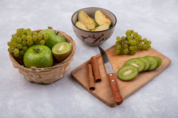 Vista lateral de rodajas de kiwi con uvas canela y un cuchillo sobre una tabla de cortar con manzanas verdes en una canasta y rodajas en un recipiente sobre un fondo blanco.