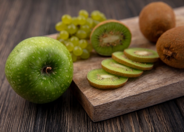 Vista lateral de rodajas de kiwi con uvas verdes en un soporte con una manzana verde sobre un fondo de madera