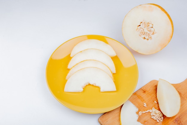 Vista lateral de rodajas de melón y semillas en placa y en la tabla de cortar con corte uno sobre fondo blanco.