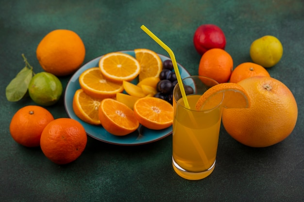 Vista lateral de rodajas de naranja con ciruela cereza sobre una placa azul con jugo de naranja pomelo y limón con lima sobre un fondo verde