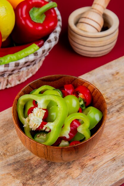 Vista lateral de rodajas de pimiento en un tazón en la tabla de cortar con verduras como tomate pimiento en la canasta con trituradora de ajo a bordo