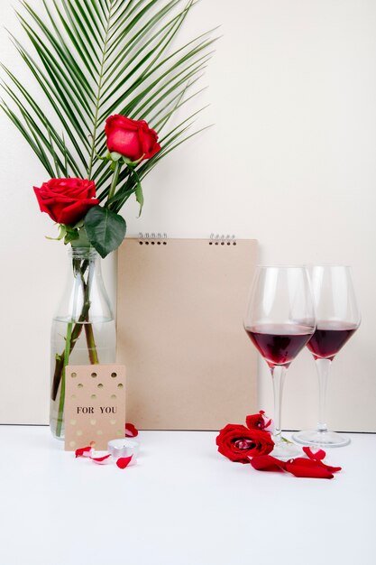 Vista lateral de rosas rojas con hoja de palma en una botella de vidrio de pie cerca de un cuaderno de bocetos y dos copas de vino tinto sobre fondo blanco.