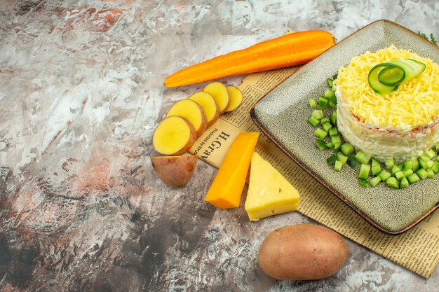 Vista lateral de una sabrosa ensalada servida con pepino picado en un periódico viejo y dos tipos de patatas con queso y zanahorias en una tabla de colores mezclados