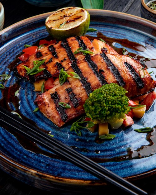 Vista lateral del salmón a la plancha con verduras, limón y salsa de soja en un plato sobre la mesa de madera