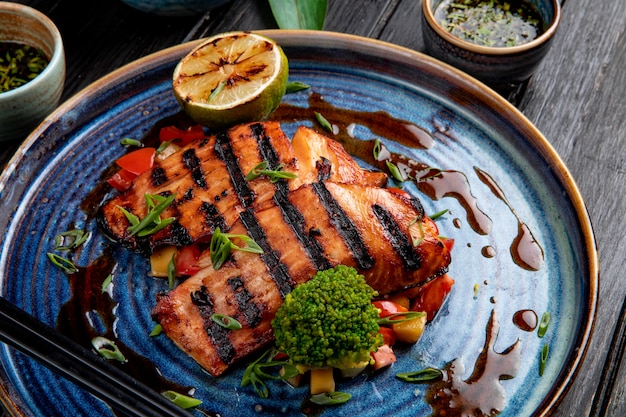 Vista lateral del salmón a la plancha con verduras, limón y salsa de soja en un plato sobre la mesa de madera