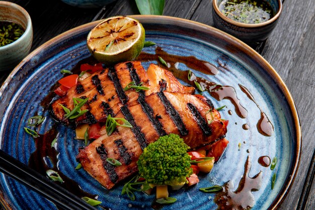 Vista lateral del salmón a la plancha con verduras, limón y salsa de soja en un plato sobre la mesa de madera