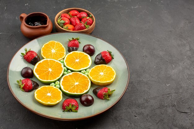 Vista lateral de salsa de chocolate y frutas, salsa de chocolate y fresas en tazones junto a caramelos de naranja y naranja picados cubiertos de chocolate sobre la mesa