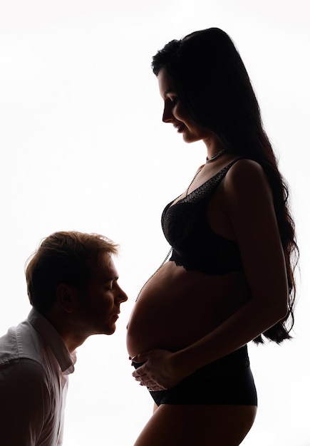 Vista lateral de la silueta del hombre marido acercándose al vientre de la mujer embarazada en elegante sujetador con cordones y bragas posando sobre fondo blanco en el estudio