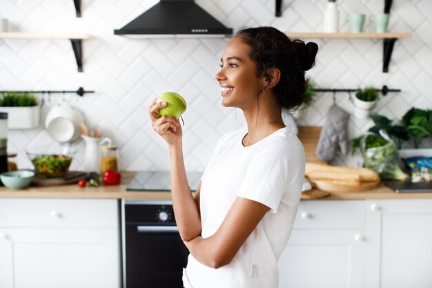 Vista lateral de la sonrisa mulata atractiva mujer que sostiene una manzana y mira lejos en la cocina blanca moderna