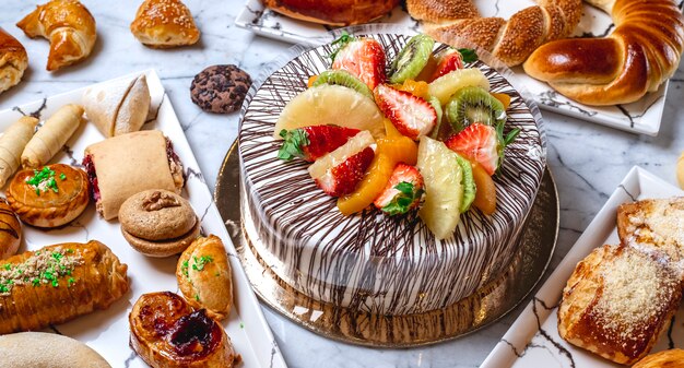 Vista lateral tarta de frutas con crema de vainilla chocolate kiwi naranja fresa piña y pasteles sobre la mesa