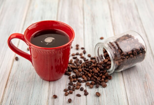 Vista lateral de la taza de café y granos de café que se derraman fuera del frasco de vidrio sobre fondo de madera