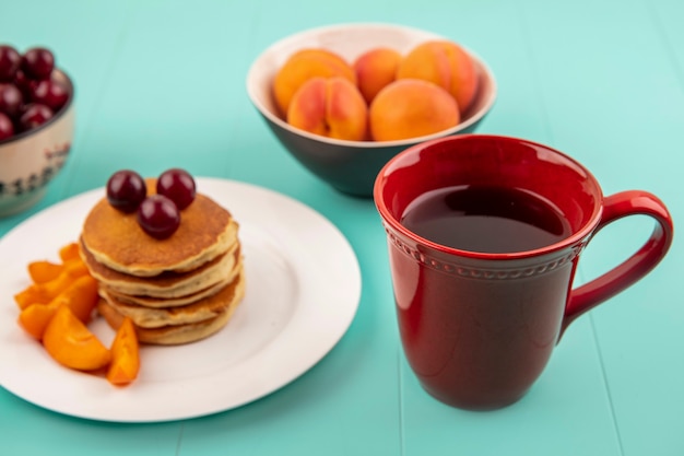 Vista lateral de la taza de café con plato de panqueques y rodajas de albaricoque con cerezas y tazones de cereza y albaricoque sobre fondo azul.