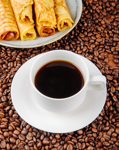 Vista lateral de una taza de café con rollo de oblea llena de leche condensada en un plato sobre granos de café