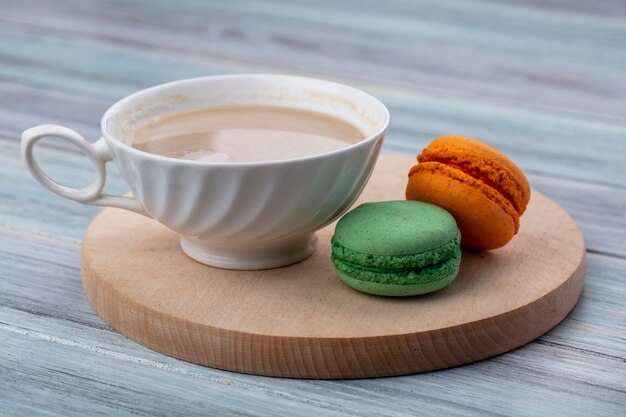 Vista lateral de la taza de capuchino con macarons de colores sobre un soporte de madera sobre una superficie gris