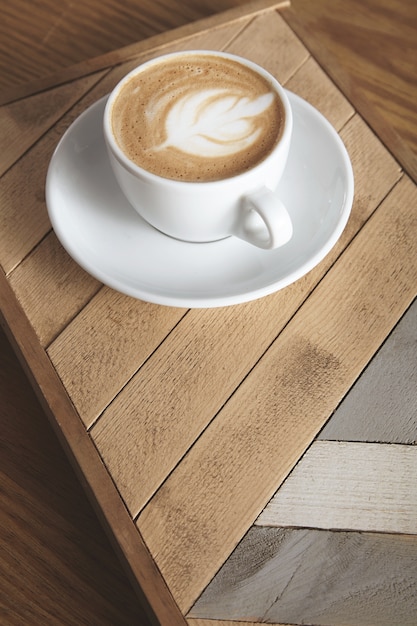 Vista lateral de la taza con cremoso capuchino latte con espuma lechosa en la parte superior en forma de hoja aislada en placa de madera con patrón. En la mesa en la presentación de la tienda de café.