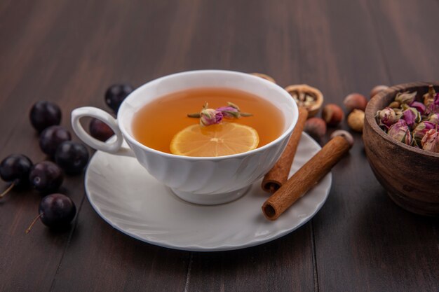 Vista lateral de la taza de hot toddy con flor de limón en el interior y canela en el platillo con endrinas, nueces y nueces y un tazón de flores sobre fondo de madera