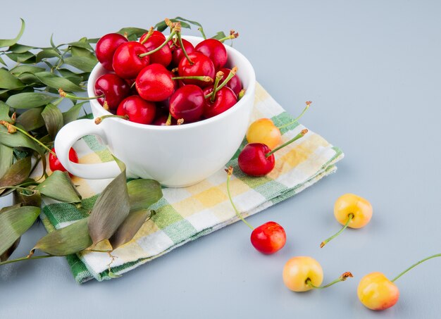 Vista lateral de la taza llena de cerezas rojas en el lado izquierdo y mesa blanca decorada con hojas