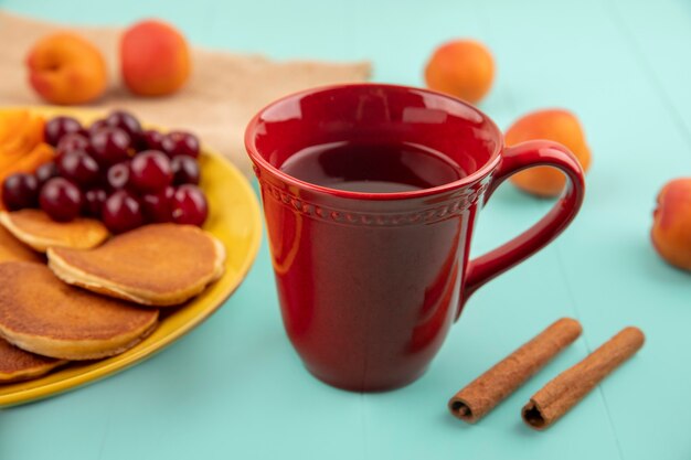 Vista lateral de la taza de té y canela y panqueques con cerezas y rodajas de albaricoque en placa y albaricoque sobre fondo azul.