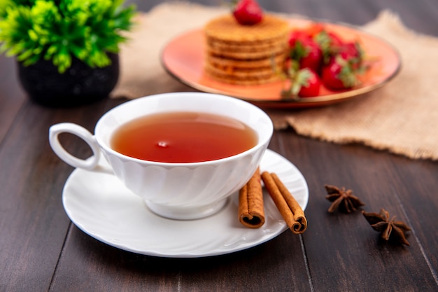 Vista lateral de la taza de té con canela en platillo y galletas waffle con fresas en placa sobre tela de saco sobre superficie de madera