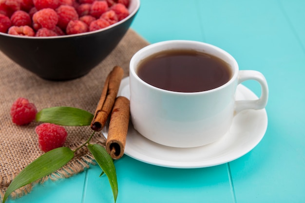 Foto gratuita vista lateral de la taza de té y canela en platillo con tazón de frambuesa y hojas de cilicio sobre superficie azul